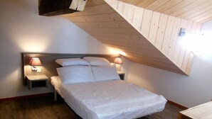 Un dormitorio con techo inclinado, una cama con ropa de cama blanca, dos mesitas de noche con lámparas, y piso de madera.