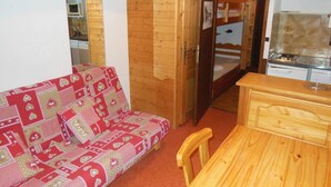 A small room with a wooden wall, a red and white patterned sofa, a wooden desk, and a kitchen area visible through an open door. - 100m from resort center, 1st floor, view village, balcony, tv, 21m², Champagny-en-Vanoise (Champagny-en-Vanoise)