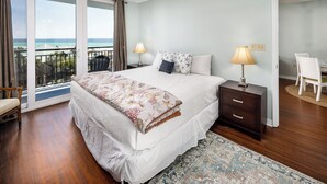 A bedroom with a large bed, a nightstand, a chair, a lamp, and a view of the ocean through a sliding door.