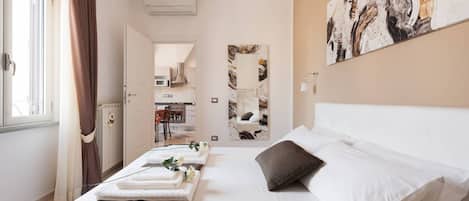 A hotel room with a bed, a window with curtains, a kitchen visible through an open door, and a large abstract painting on the wall.