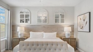 Een slaapkamer met een groot bed, twee nachtkastjes met lampen, een gestoffeerde fauteuil en drie rondbogige ramen met kroonluchters.