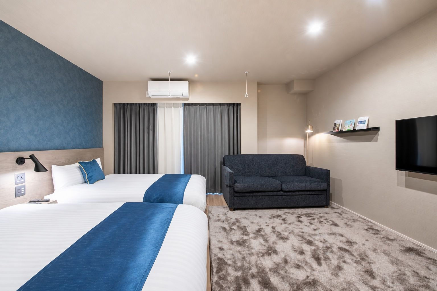 A hotel room with two beds, a sofa, a flat-screen TV, and a wall-mounted shelf.