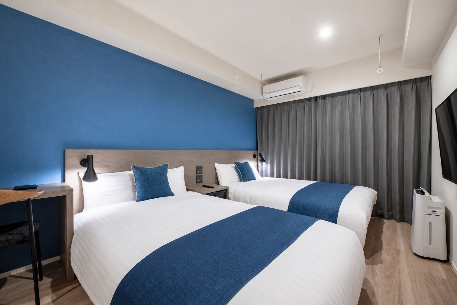 A hotel room with two beds, a desk, a lamp, an air conditioning unit, and a TV.