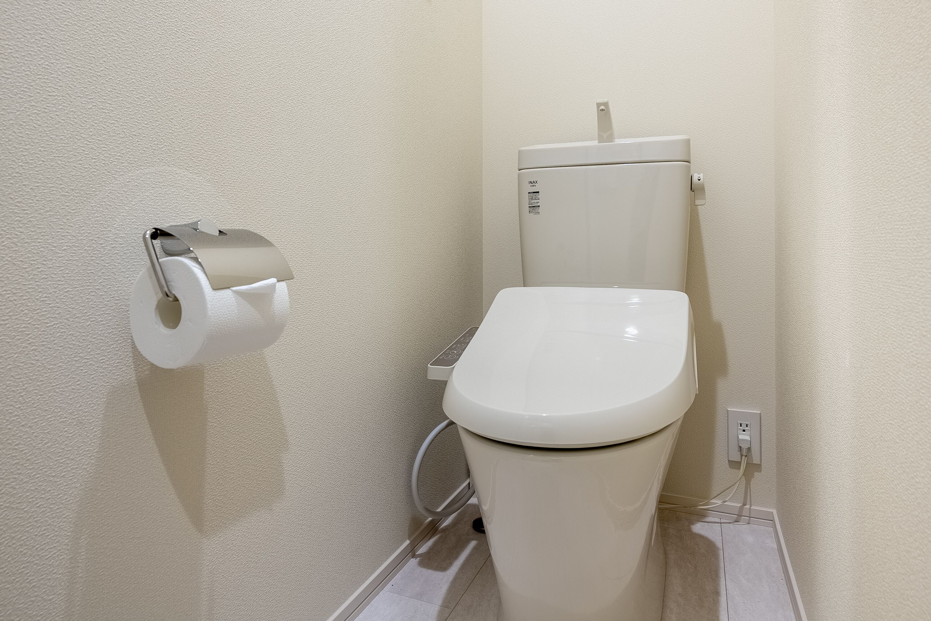 A toilet with a paper towel holder, a visible water pipe, and a wall-mounted electrical outlet.