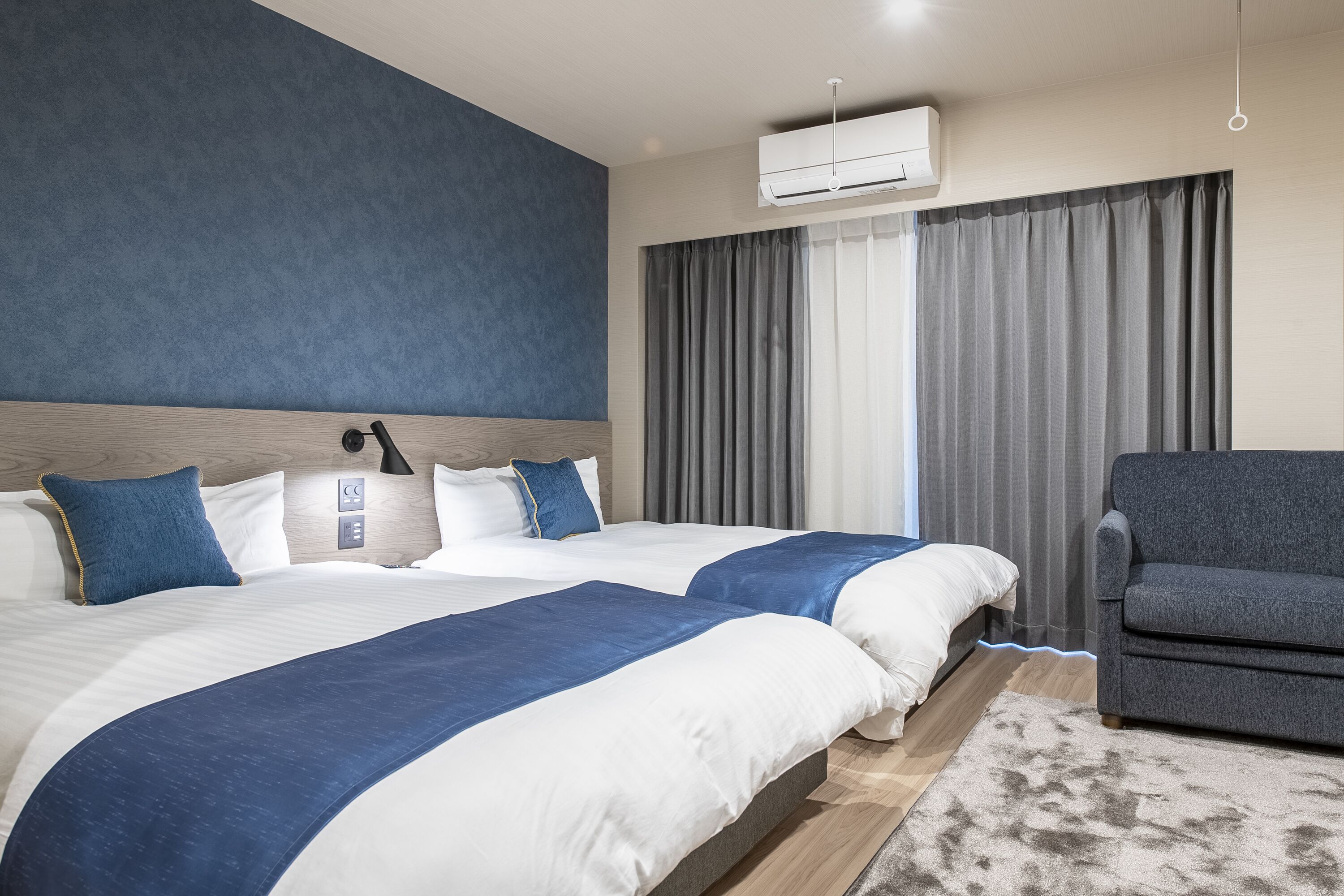 A hotel room with two beds, a grey sofa, a large window with grey curtains, and an air conditioning unit.