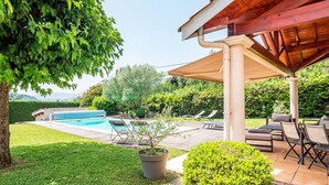 Zona de piscina con un porche cubierto, mobiliario y vistas al paisaje.