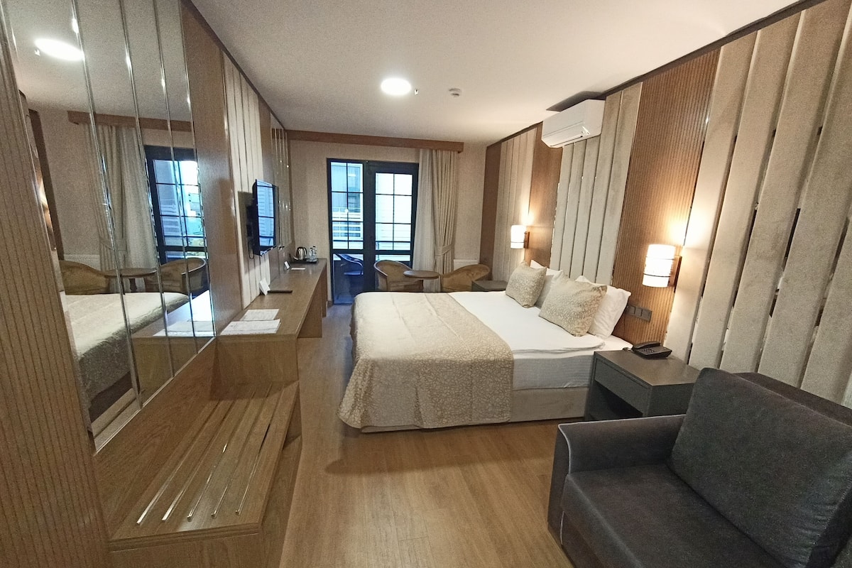 A hotel room with a large bed, a sofa, a desk with a chair, a TV, and a window with curtains.