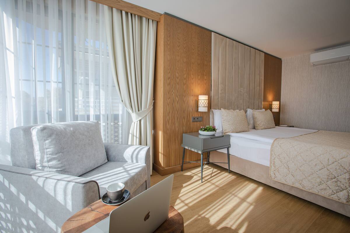 A hotel room with a sofa, a bed, a small table with a lamp, and a laptop on a wooden side table.