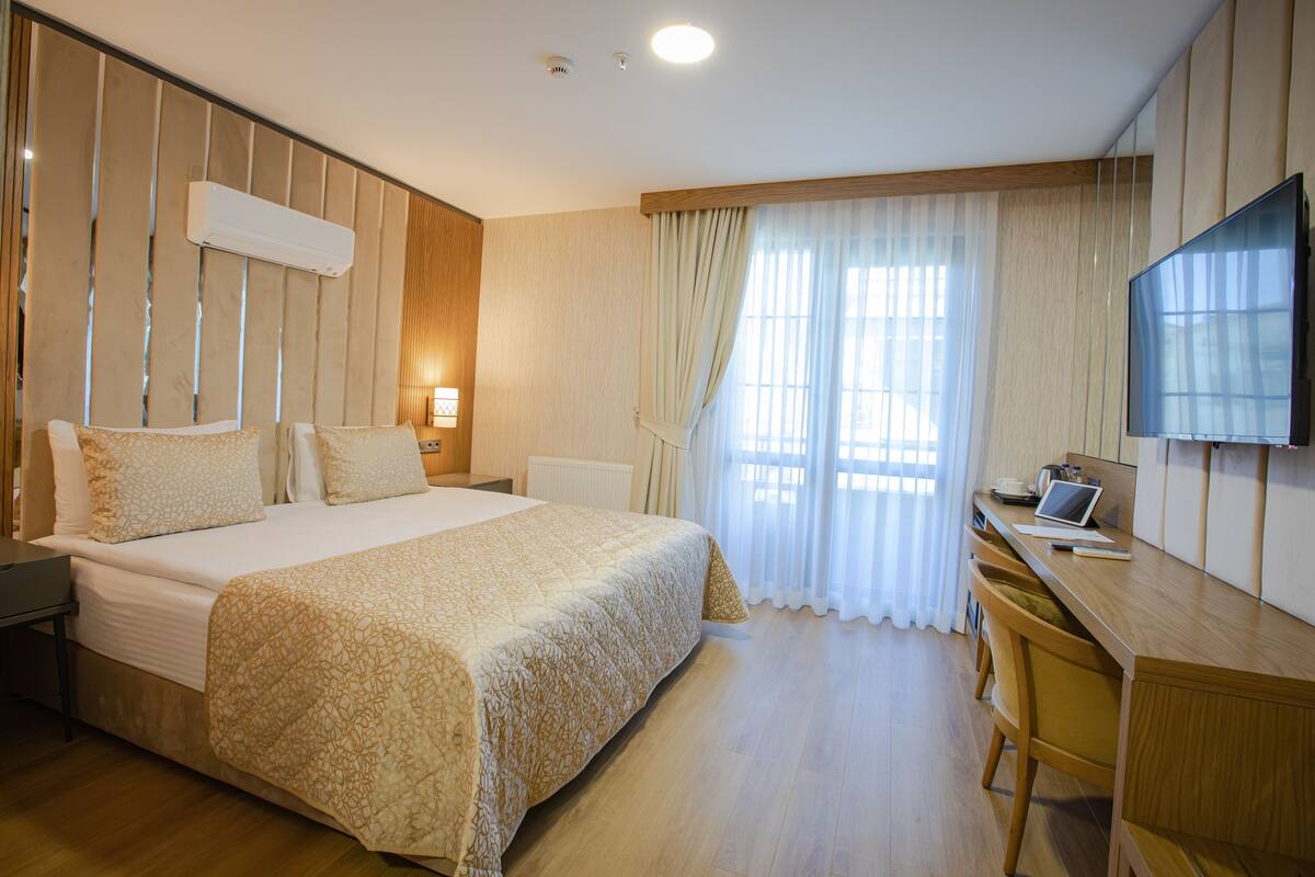 A hotel room with a large bed, a desk with a laptop, a flat-screen TV, and a window with curtains.