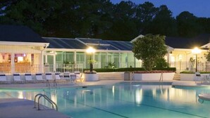 Ein Swimmingpool mit Liegestühlen und Barbereich, umgeben von Bäumen und Gebäuden mit großen Fenstern.