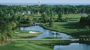 Ein Golfplatz mit einem Wasserhindernis, Sandbunkern und einem Wasserturm im Hintergrund.