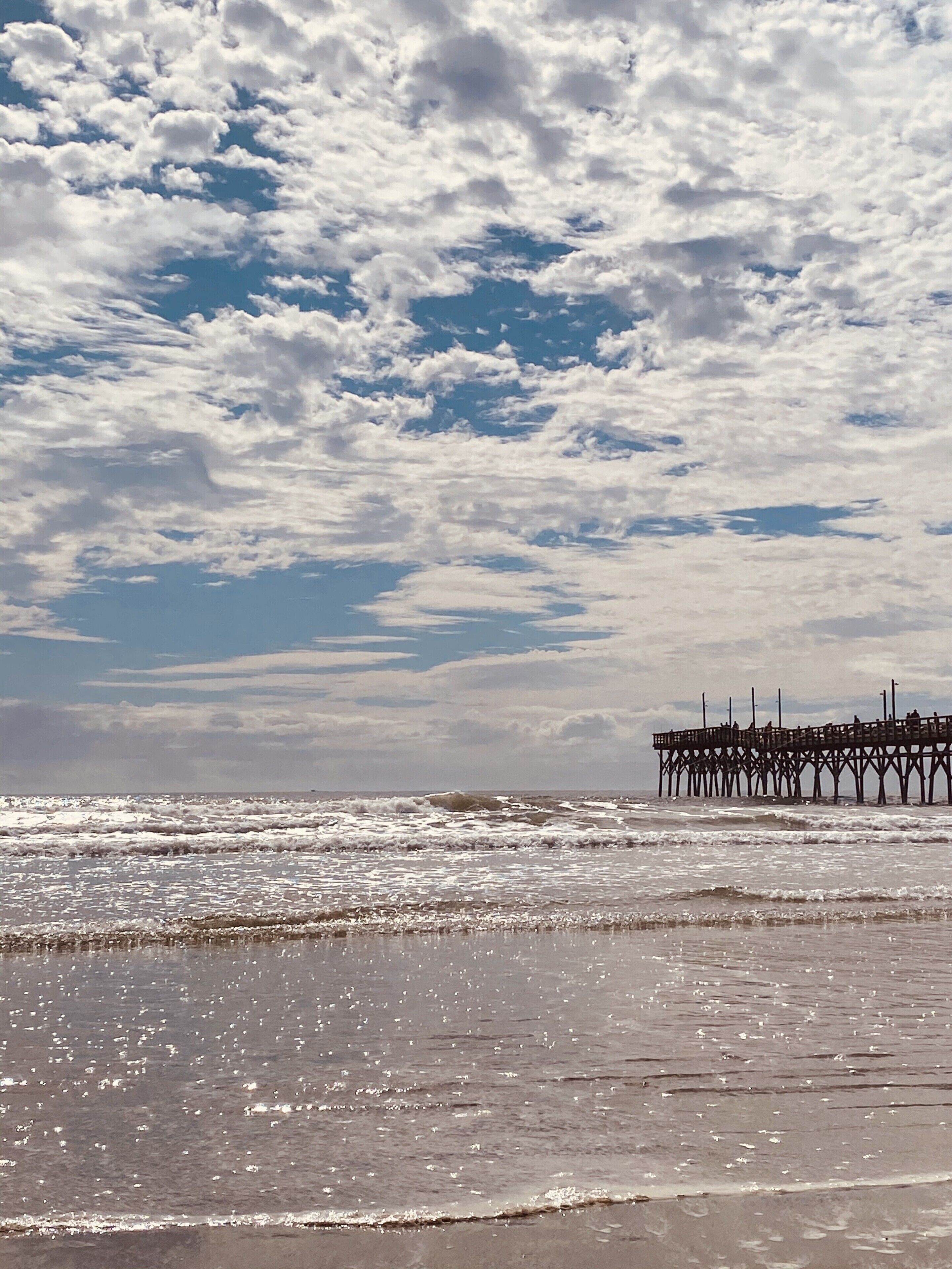 A beach with waves, a pier, and a partly cloudy sky.