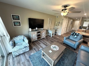 A living room with a flat-screen TV, a coffee table, a sofa, and a chair. There is a ceiling fan, a bookshelf, and a dining area in the background.