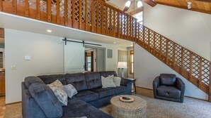 A spacious living room with a large sectional sofa, a small round coffee table, and a single armchair. A wooden staircase with a lattice design leads to an upper level. - PNRD11 - Updated Home, AC, SHARC Passes, Private Hot Tub (Sunriver)