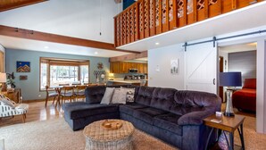 A spacious living room with a dark sectional sofa, a round wicker ottoman, and a dining area with wooden chairs and a table. - PNRD11 - Updated Home, AC, SHARC Passes, Private Hot Tub (Sunriver)