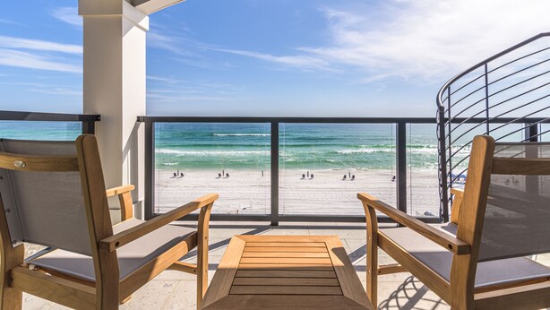 A balcony with wooden chairs and a view of the beach and ocean.