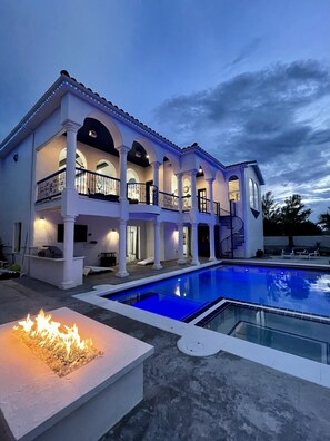 A large, multi-story house with a swimming pool, outdoor lighting, and a fire feature.