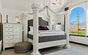 A four-poster bed with a dark headboard, a white cabinet with drawers, a round ottoman, and a view of mountains through a window.