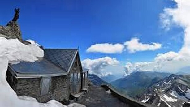 Un chalet de montagne aux murs en pierre et au toit en pente, entouré de neige et de montagnes.
