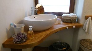 A bathroom with a large white sink, a wooden vanity, a small trash bin, and a wicker basket. - House Typical Of The Susa Valley (San Giorio di Susa)