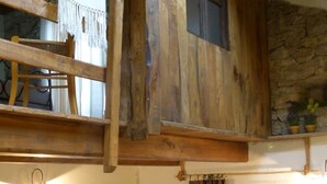 Une pièce rustique avec un plafond en bois, un espace en mezzanine doté d’un fauteuil, une porte avec un panier, et une fenêtre avec des rideaux.