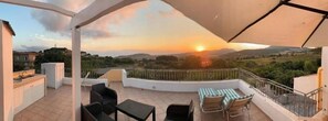 Eine Terrasse mit Blick auf den Sonnenuntergang, Gartenmöbel und einer kleinen Küchenzeile.