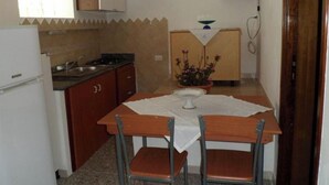 A small kitchen with a refrigerator, stove, sink, and wooden cabinets. There is a table with two chairs and a potted plant on it. - Mare Natura (Castelsardo)