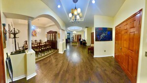 A spacious hallway with a dining area, wooden flooring, a chandelier, and a large painting on the wall.