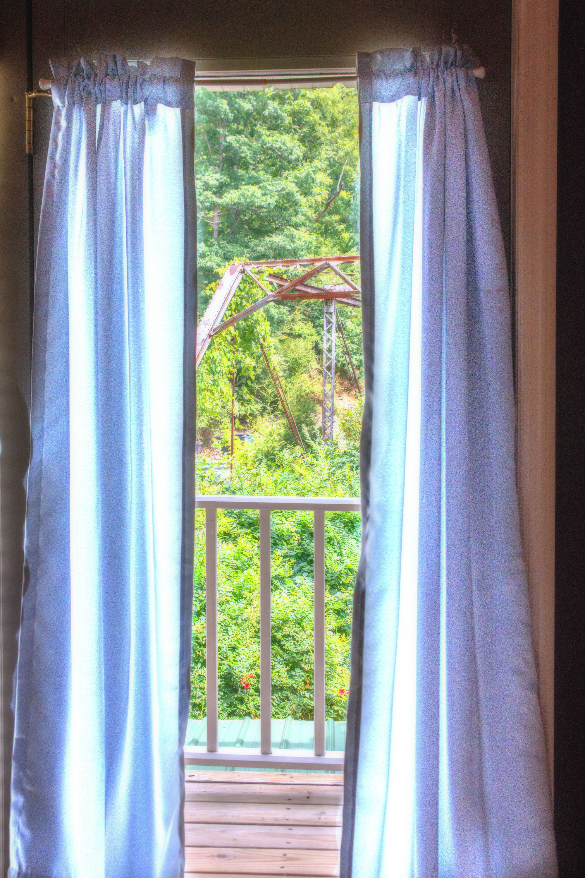 Vista a través de una ventana con cortinas blancas, mostrando una terraza de madera y abundante vegetación en el exterior.