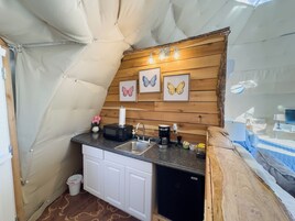 Cocina compacta con lavabo, microondas y cafetera. En la pared hay cuadros con mariposas y un pequeño cubo de basura.