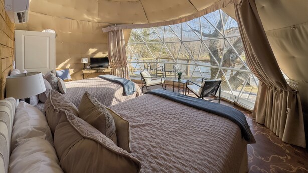 Une pièce confortable, de style tente, bien éclairée, avec un grand lit, un bureau doté d’une lampe et une vue sur un plan d’eau grâce à de grandes fenêtres.