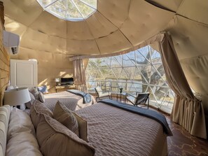 Une pièce confortable, de style tente, bien éclairée, avec un grand lit, un bureau doté d’une lampe et une vue sur un plan d’eau grâce à de grandes fenêtres.