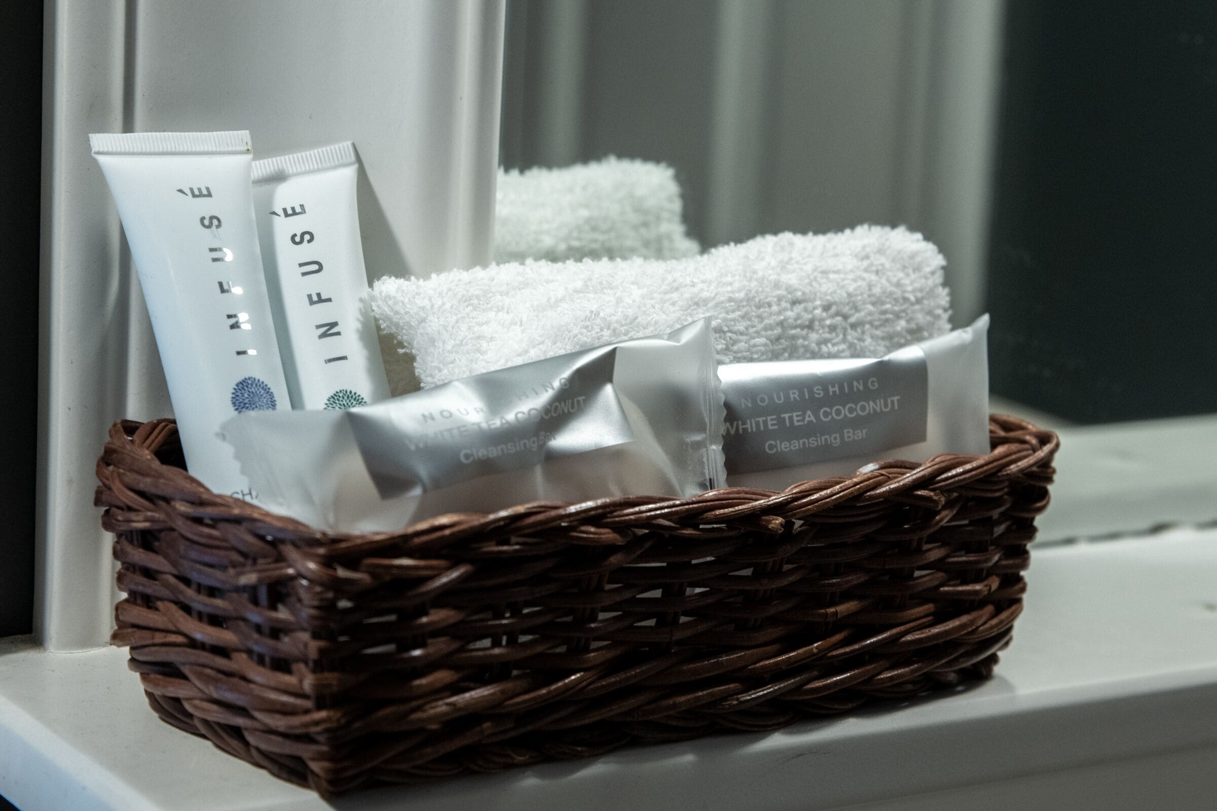 Un panier en osier contenant des serviettes blanches et deux tubes de produits de soins pour la peau, posé sur un rebord de fenêtre.
