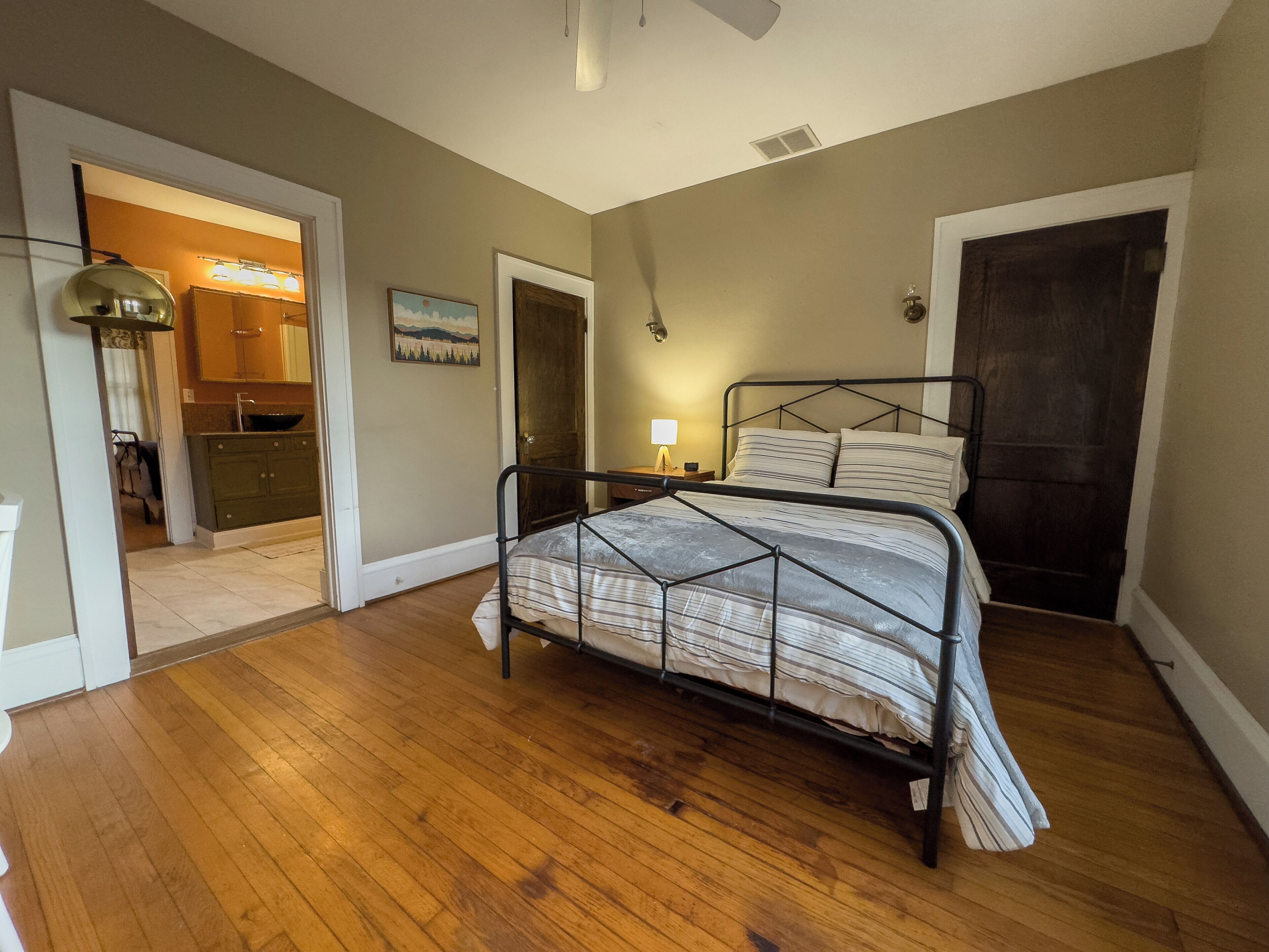 Un dormitorio con estructura de cama metálica, suelo de madera, mesita de noche con lámpara y puerta que da al baño.