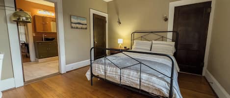 Un dormitorio con estructura de cama metálica, suelo de madera, mesita de noche con lámpara y puerta que da al baño.