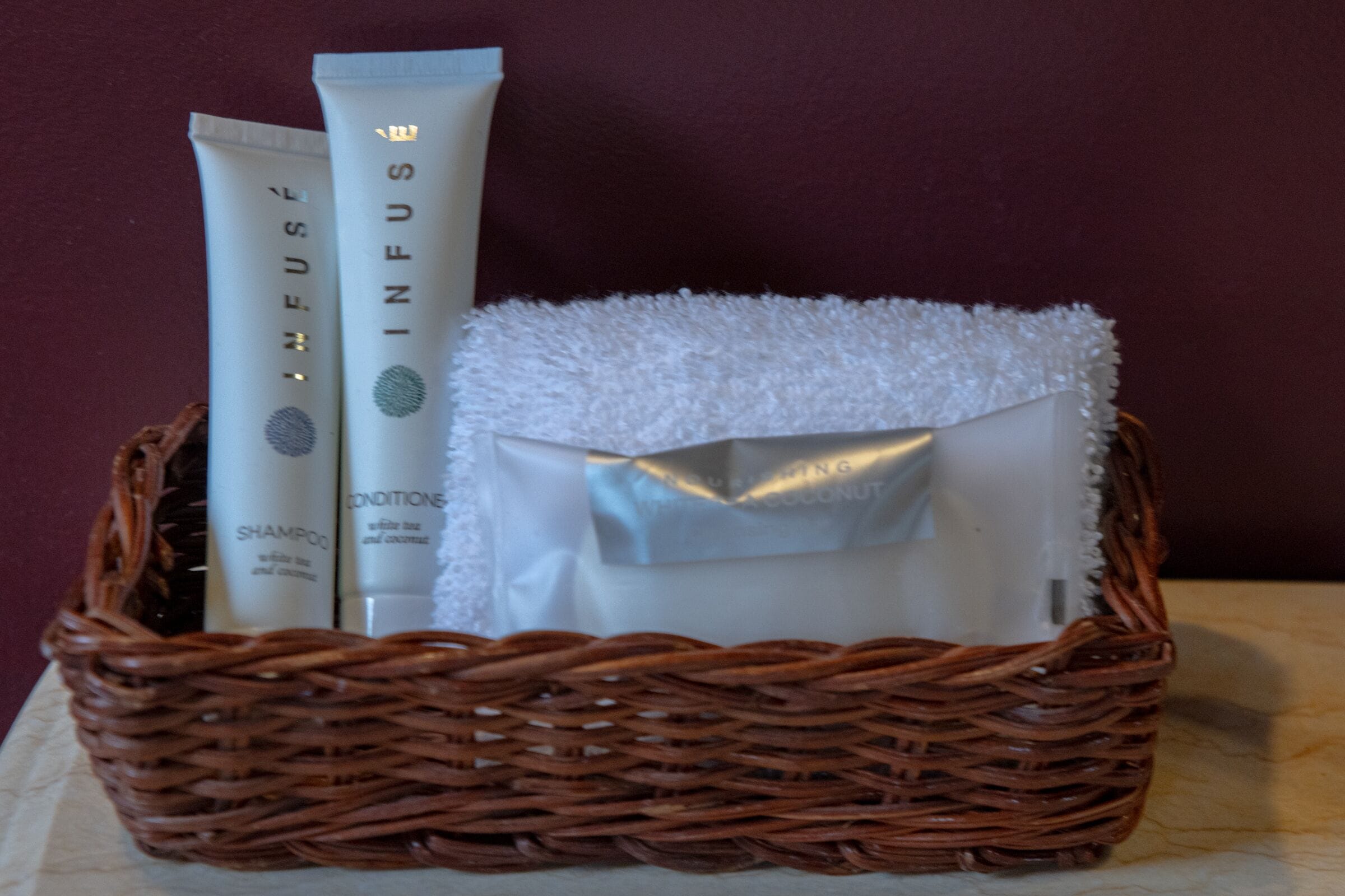 A wicker basket containing a white towel, a tube of shampoo, and a tube of conditioner.