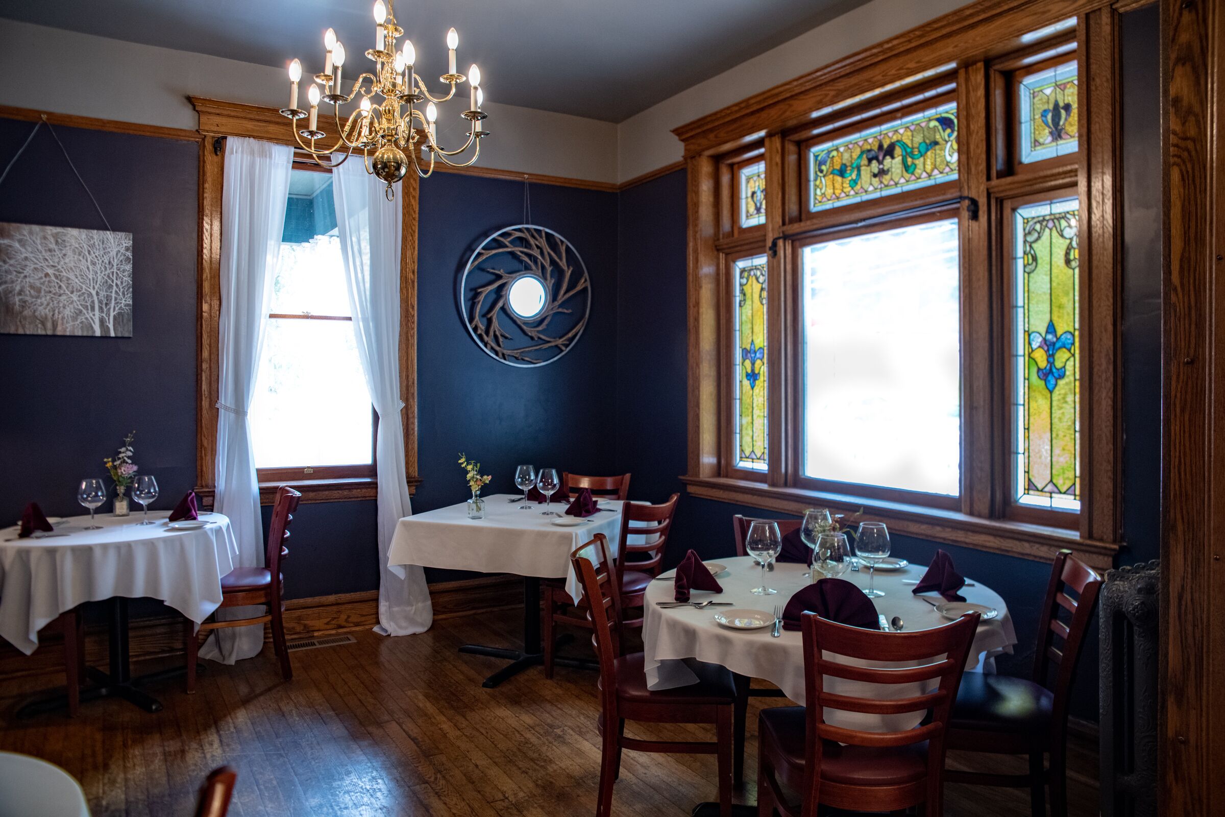 A dining area with wooden tables and chairs, a chandelier, stained glass windows, and a wall-mounted circular decoration.