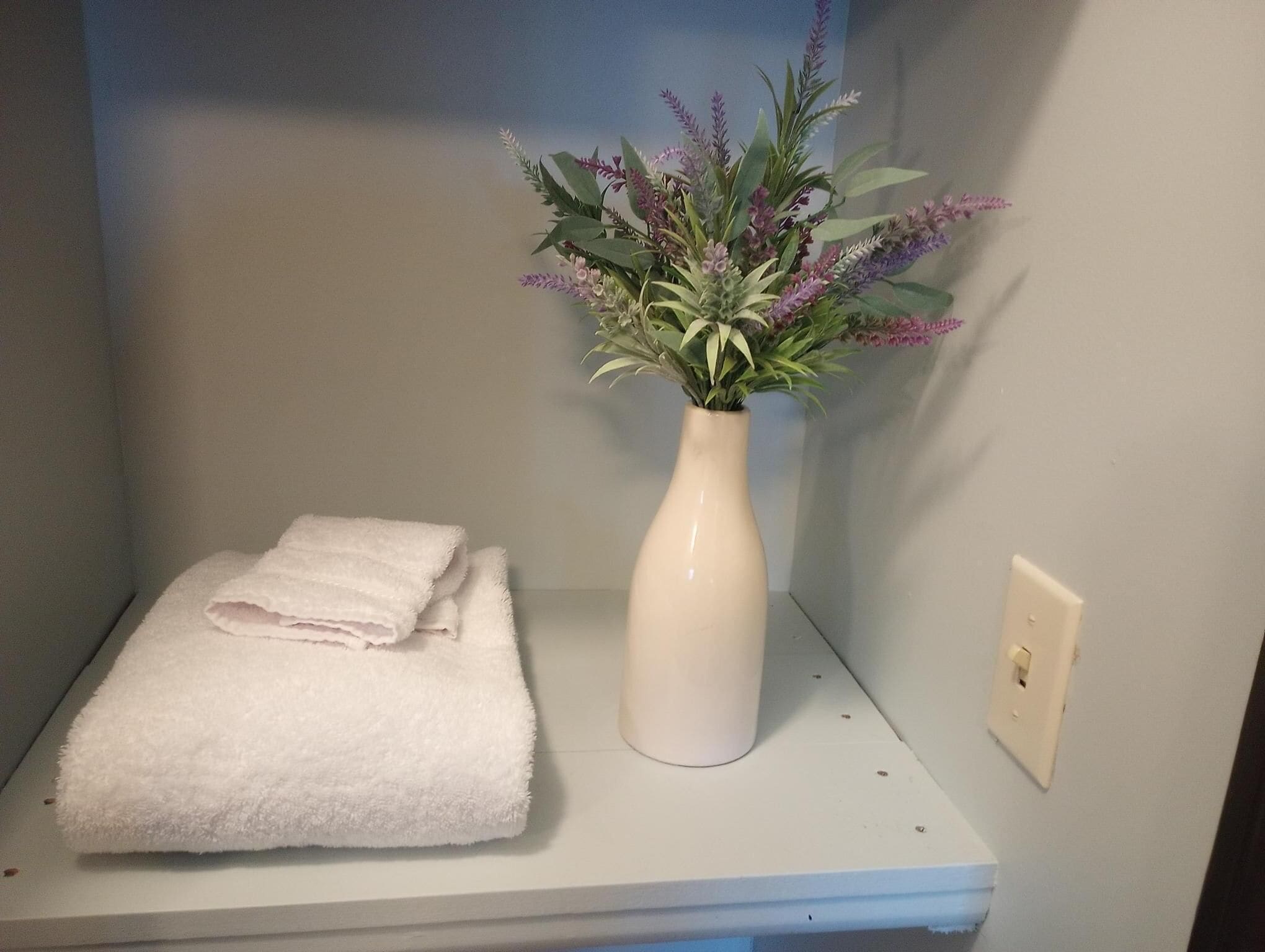 A bathroom with a white towel folded on a shelf and a white vase with purple flowers.