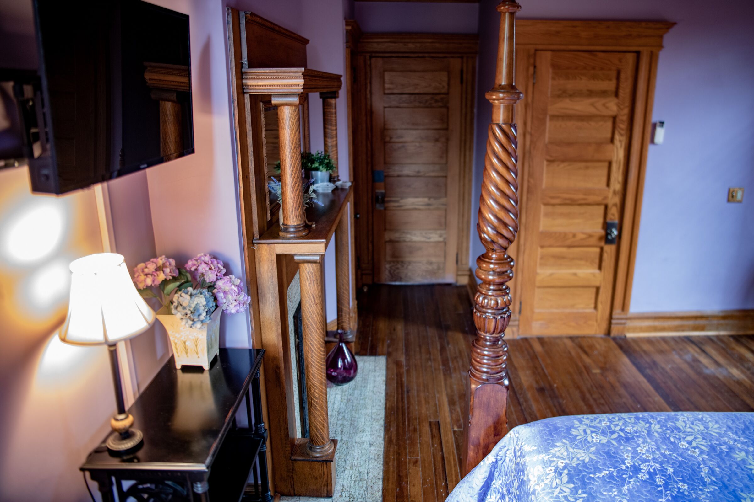 Una habitación con piso de madera, una cama con colcha azul estampada de flores, una silla de madera, una mesa con lámpara y un espejo.