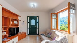 Un salon avec un téléviseur, des étagères en bois, une porte verte, un canapé beige et une fenêtre donnant sur un beau paysage.