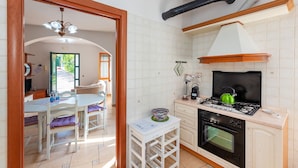 Une cuisine compacte équipée d’une cuisinière à gaz, d’un four et d’une bouilloire verte. On y trouve une petite table avec des chaises et une fenêtre donnant sur un jardin.