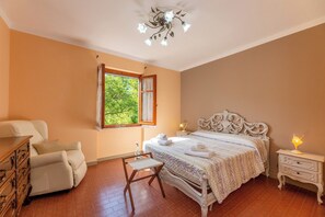 Een slaapkamer met een groot bed, een dressoir, een stoel, een nachtkastje en een raam met uitzicht op groen.