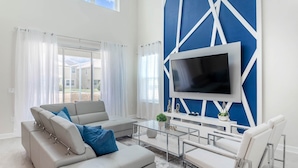 Una sala de estar moderna con un televisor de pantalla plana grande montado en una pared azul decorativa, un sofá color beige con cojines azules, una mesa de centro de vidrio y un sillón blanco.
