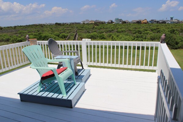 A deck with two Adirondack chairs and a small table, overlooking a grassy area with houses in the background.