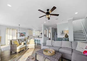 A spacious living area with a grey sofa, wicker furniture, a ceiling fan, and a kitchen in the background.
