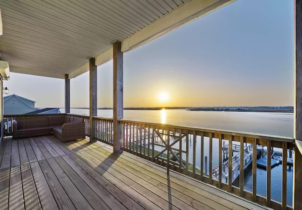 A wooden deck with a view of a sunset over water, a railing, and a bench.