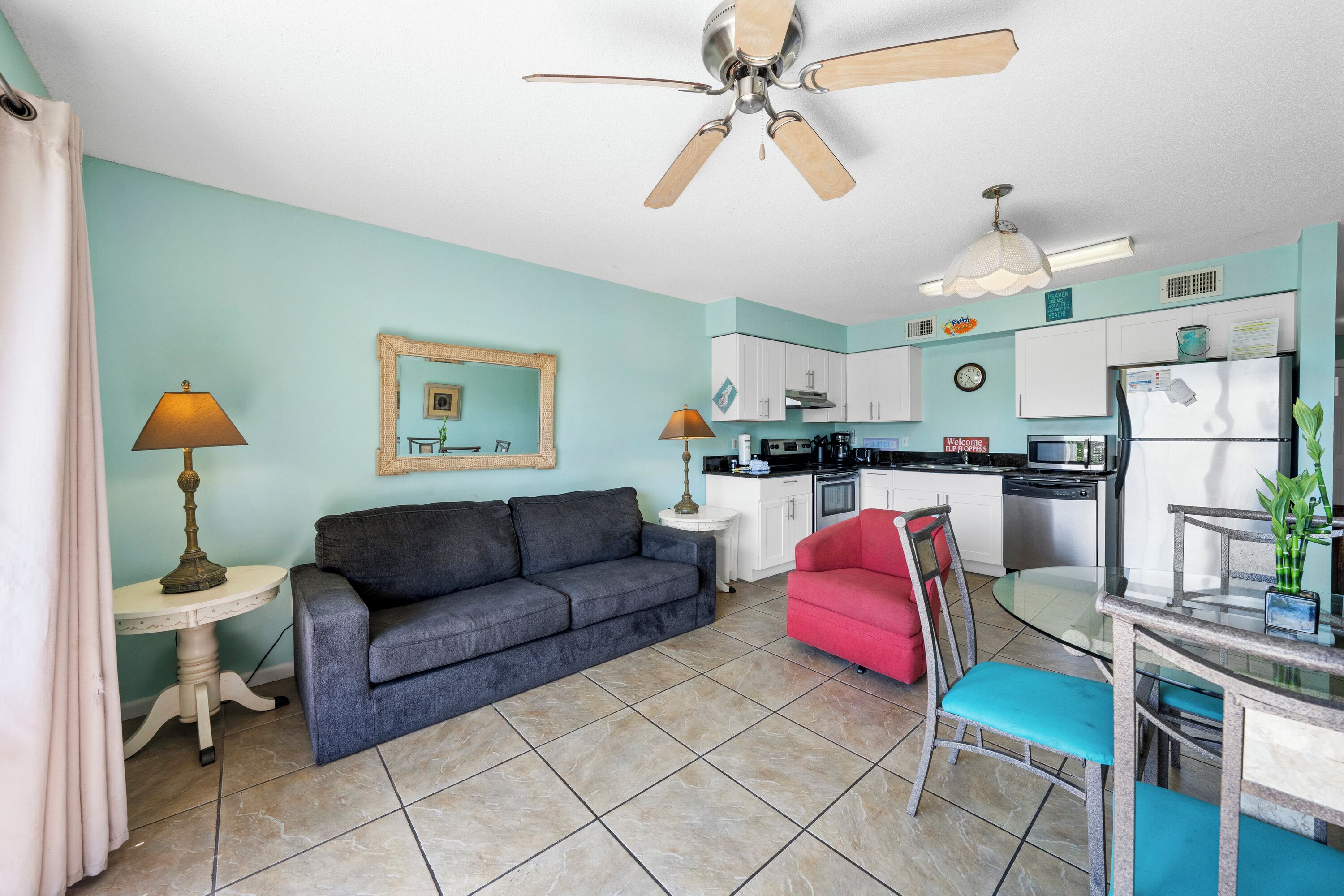 A living room with a sofa, a red chair, a glass table, and a kitchen area with cabinets and appliances.