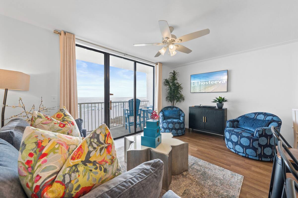 A modern living room with a sofa, armchairs, a ceiling fan, a balcony with a view, and a plant.