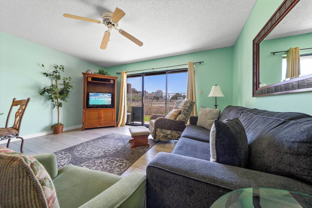 A living room with a sofa, armchairs, a TV, a ceiling fan, a plant, and a view through a glass door.