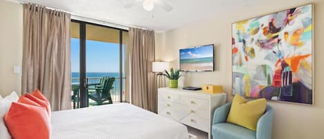 A hotel room with a bed, a desk, a chair, a ceiling fan, a TV, a painting, and a view of the ocean.